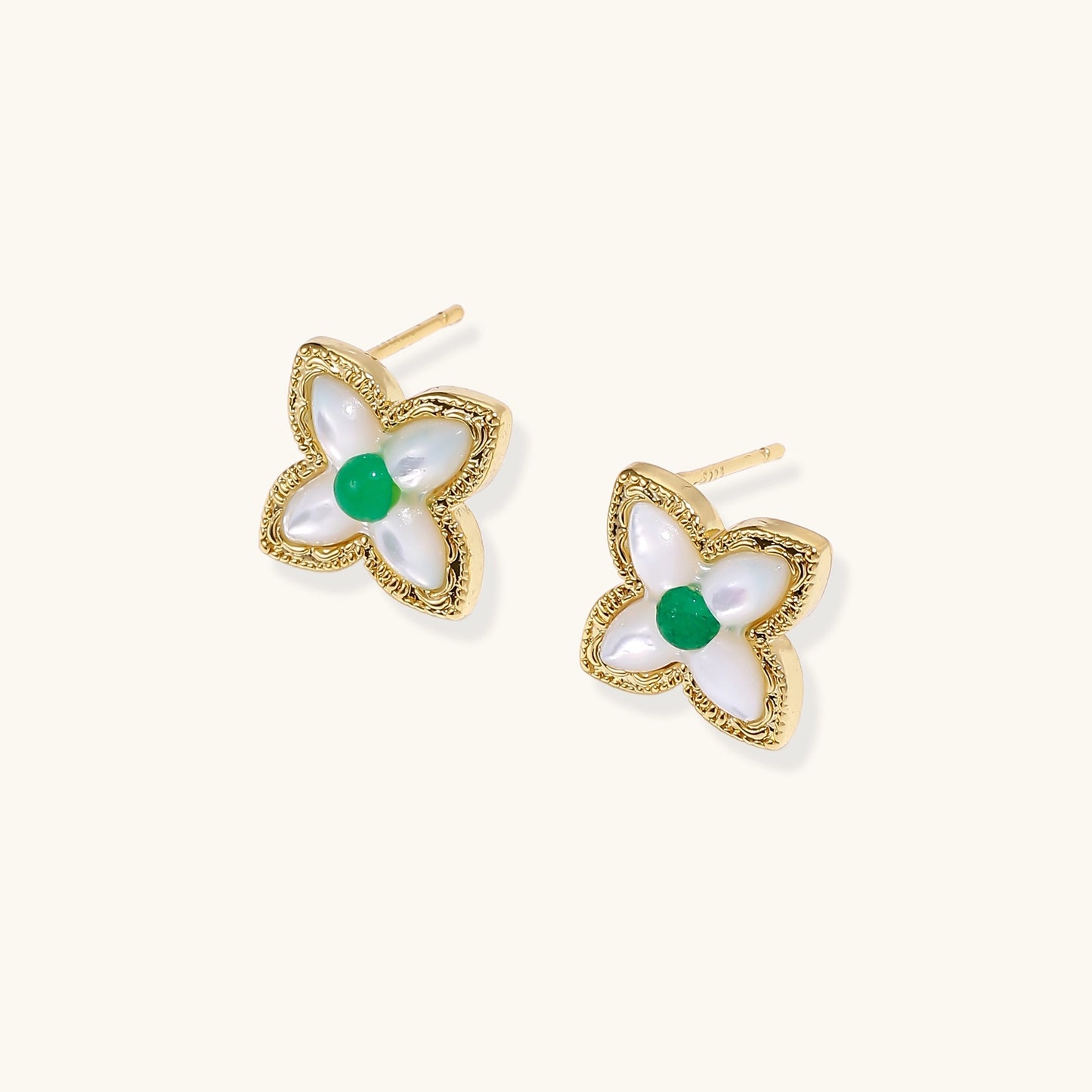 Dainty gold-plated quatrefoil stud earrings with white mother-of-pearl petals and green gemstone centers on beige background.