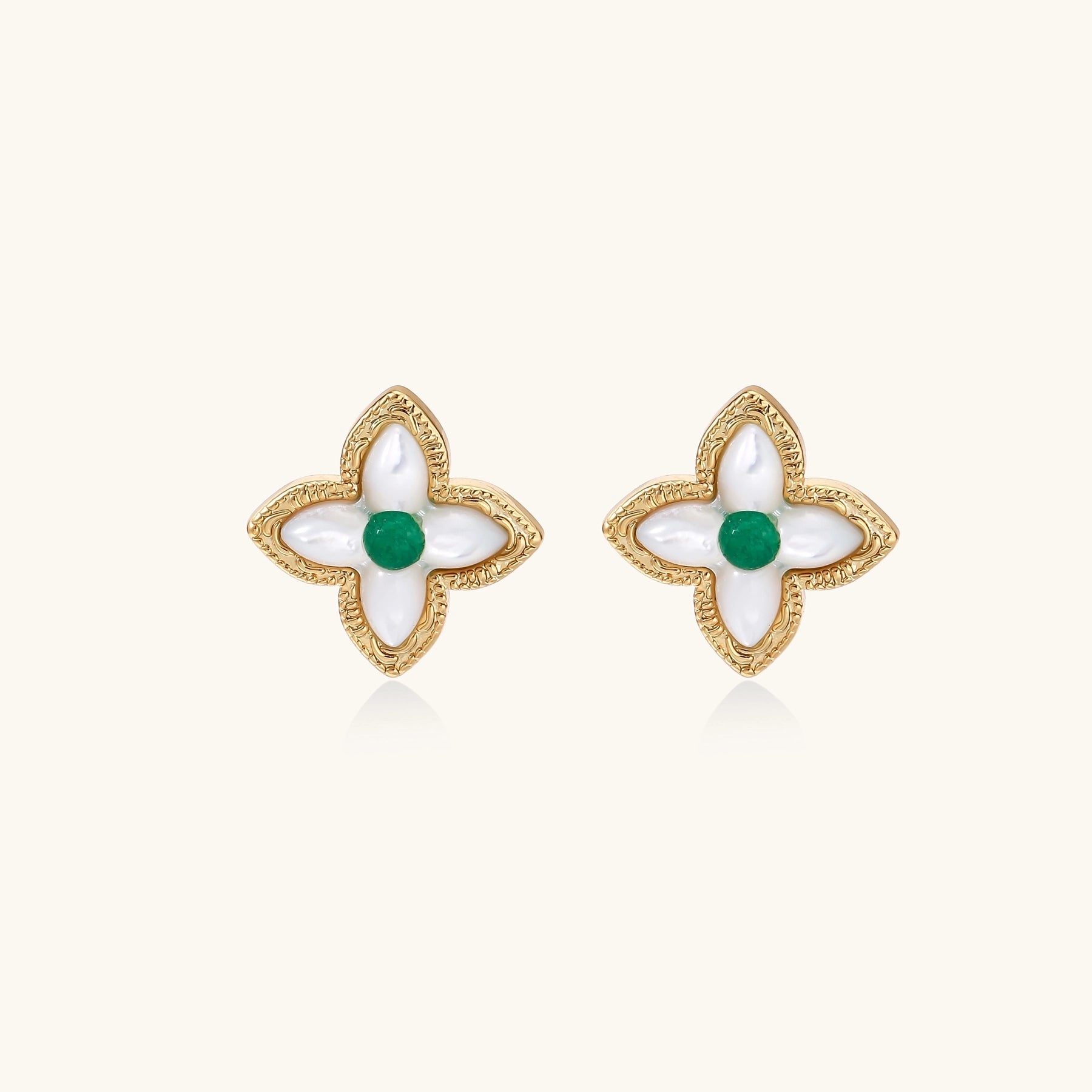 Dainty gold-plated quatrefoil stud earrings with white mother-of-pearl petals and green gemstone centers on beige background.